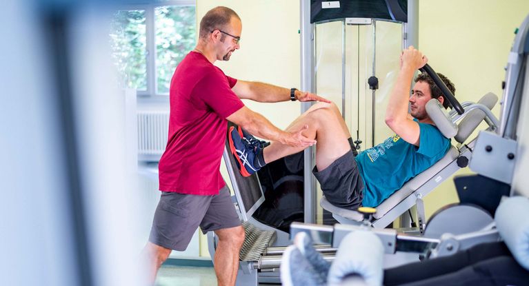 Gerätetraining – Orthopädische Reha Therapeut unterstützt Patienten beim Gerätetraining im MTT-Raum
